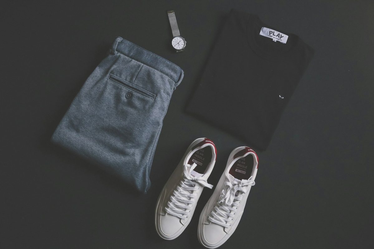 Flat lay of a black shirt, grey trousers, a watch and white trainers on a dark surface